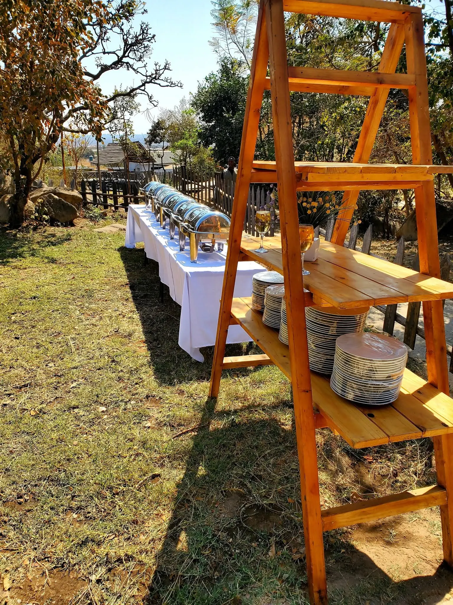 Outdoor buffet table with chafing dishes and a wooden plate shelf on the lawn