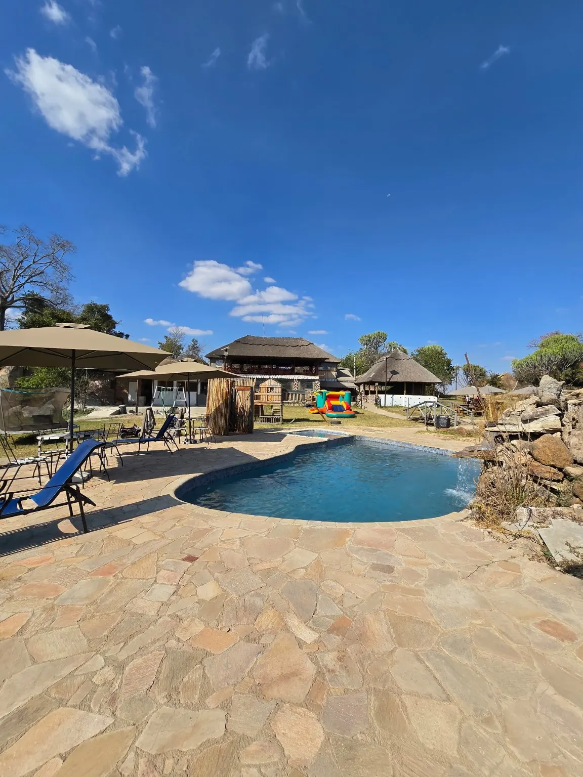 Resort swimming pool with sun loungers, umbrellas and thatched buildings under a blue sky