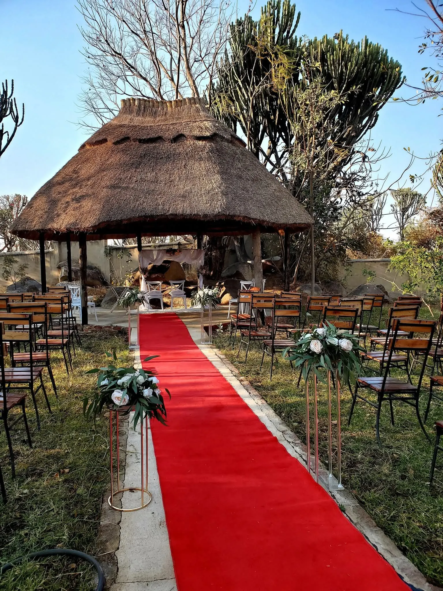 Outdoor ceremony set-up: red carpet aisle, chairs and flower stands leading to a thatched gazebo