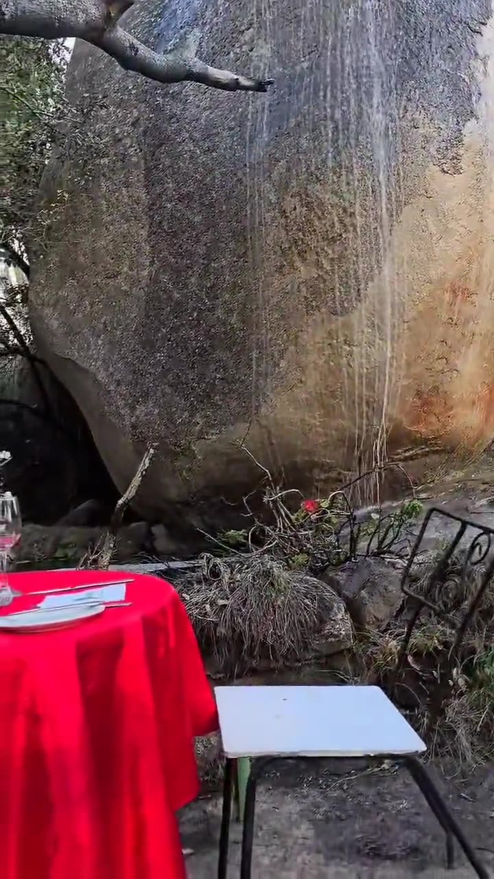A table dressed in red with glasses and flowers set among large boulders and trees